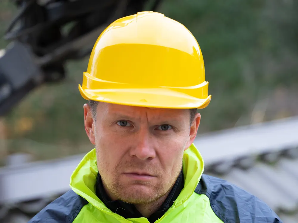 Tekijä kypärässä ja huomioliivissä työmaalla, takana kaivinkoneen puomi